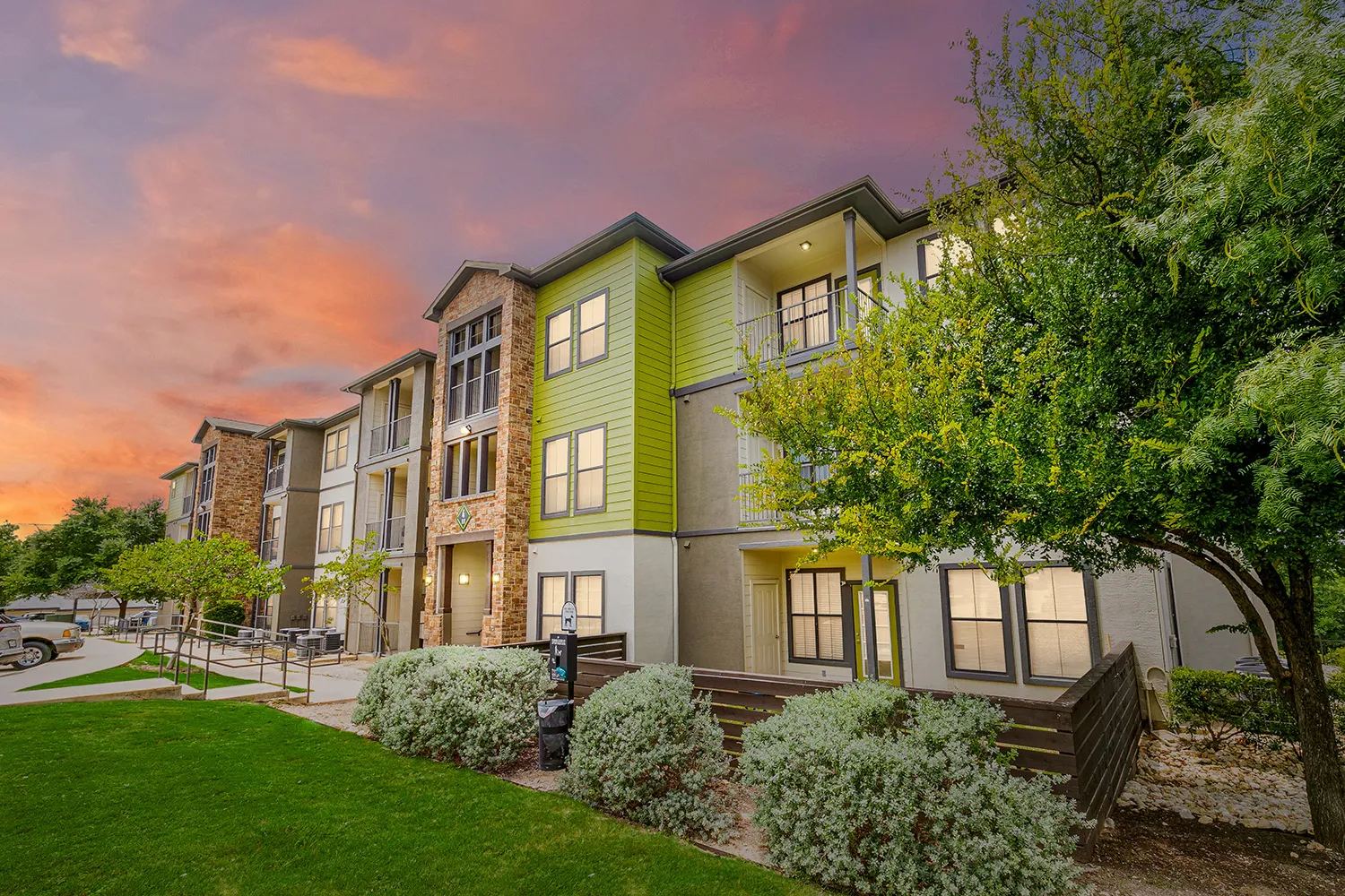 The Benton apartment building with green siding and brick surrounded by landscaping at dusk. 